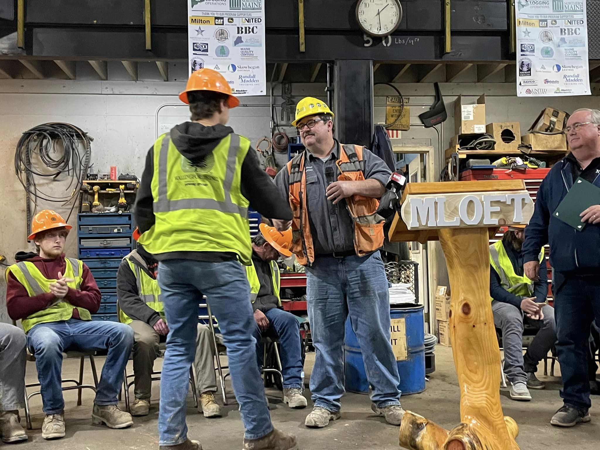 photo of two people in safety vests and hard hats shaking hands in a warehouse with a podium that reads "MLOFT"