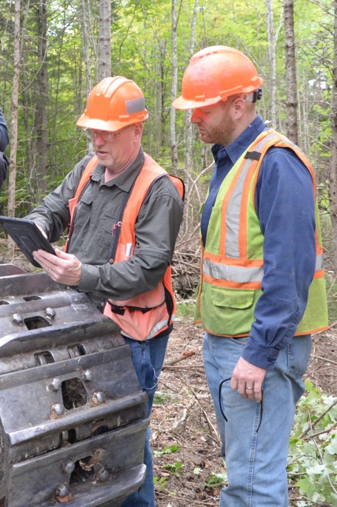 photo of two men in safety vests and hardhats talking over a tablet in a forest next to logging equipment