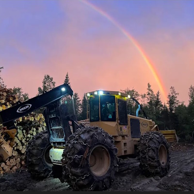 photo of logging equipment in front of a stack of logs with a rainbow in the background