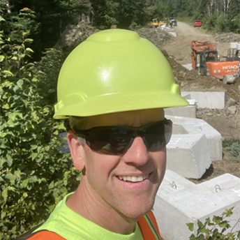 A selfie of a smiling construction worker wearing a high-visibility lime green hard hat, dark sunglasses, a lime green shirt, and an orange safety vest with reflective silver stripes. He is standing outdoors at a construction site located in a wooded, mountainous area. In the background, there is an orange Hitachi excavator, several large concrete blocks, a dirt road, and lush green trees under a clear blue sky.